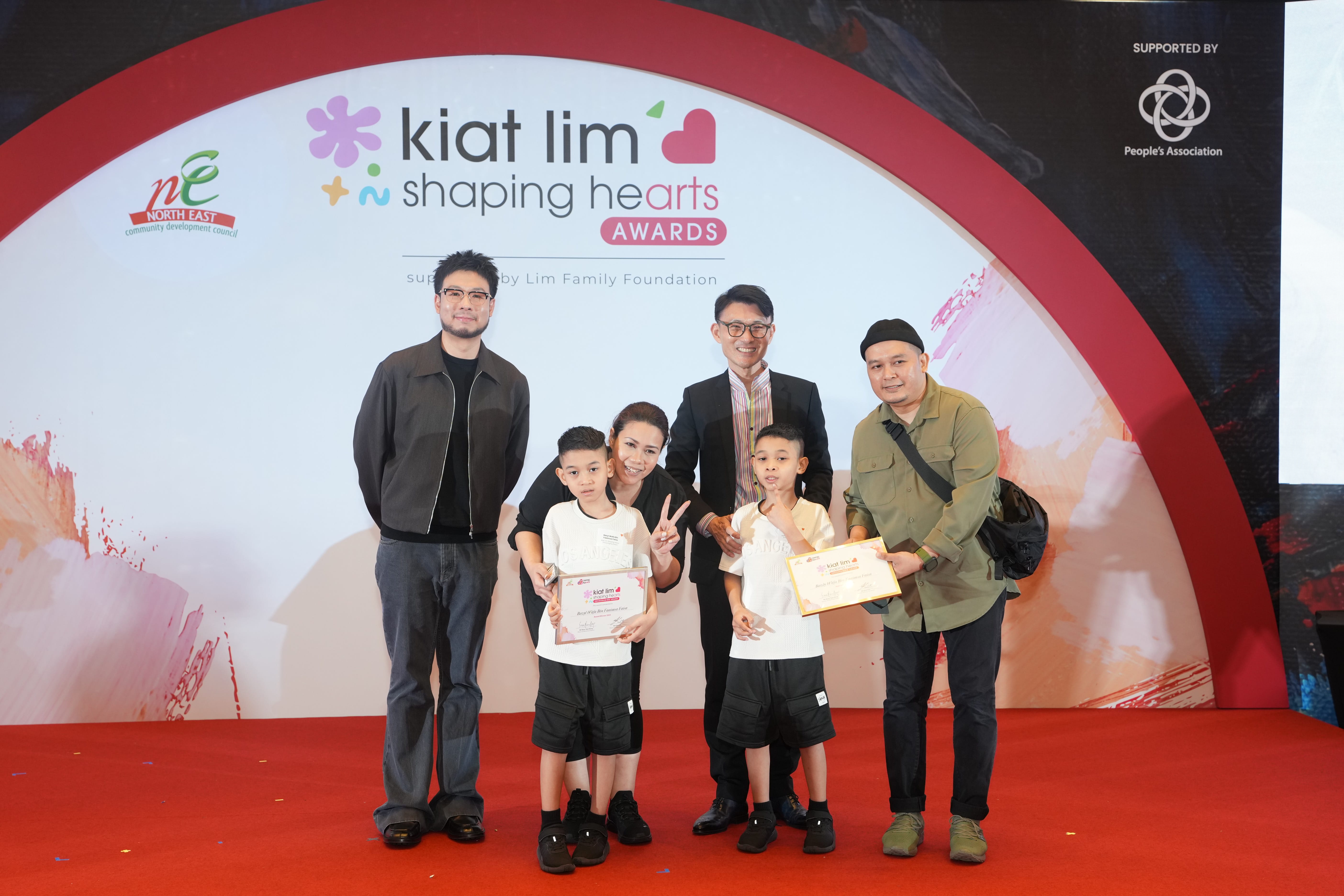 Award recipient standing on stage with the presenter, holding their certificate and posing for a group photo during the award ceremony, with a backdrop reading ‘Kiat Lim-Shaping Hearts Awards'.